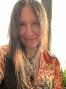 Smiling brown-haired woman with gray in her hair standing in front of a window in an orange paisley dress.