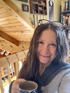 Middle-aged woman with brown hair in a grey sweater standing in front of a bookcase and collection of art framed on the wall.